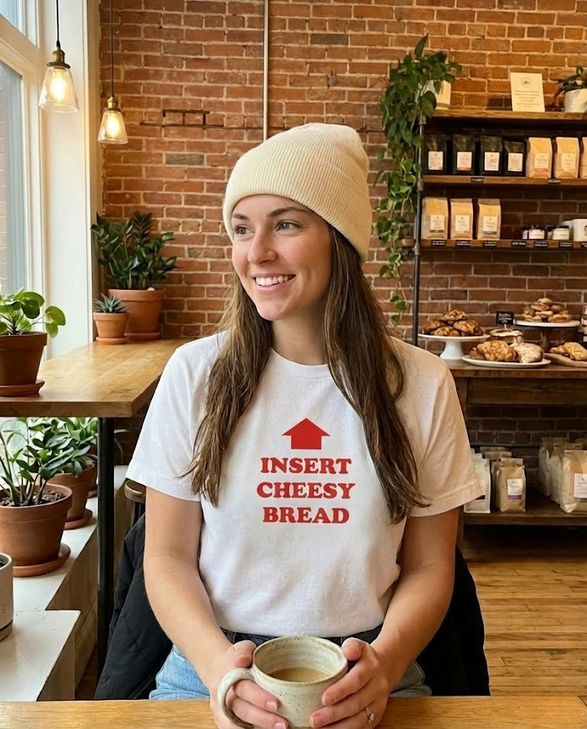 Woman wearing a white t-shirt with red text "Insert Cheesy Bread" in a cozy cafe setting